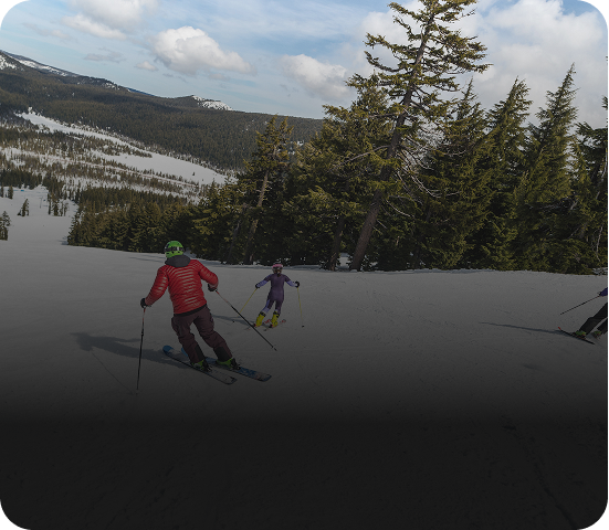 Two skiers descending a snow-covered slope with trees and mountains in the background.